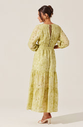 A woman is wearing a long, yellow dress made of lightweight, embroidered fabric with eyelet details. The dress features long puffed sleeves, a gathered waist, and a tiered skirt that reaches her ankles. The back of the dress has a keyhole opening tied with a small bow at the neck. She is also wearing beige high-heeled shoes and has her hair styled in an elegant updo.